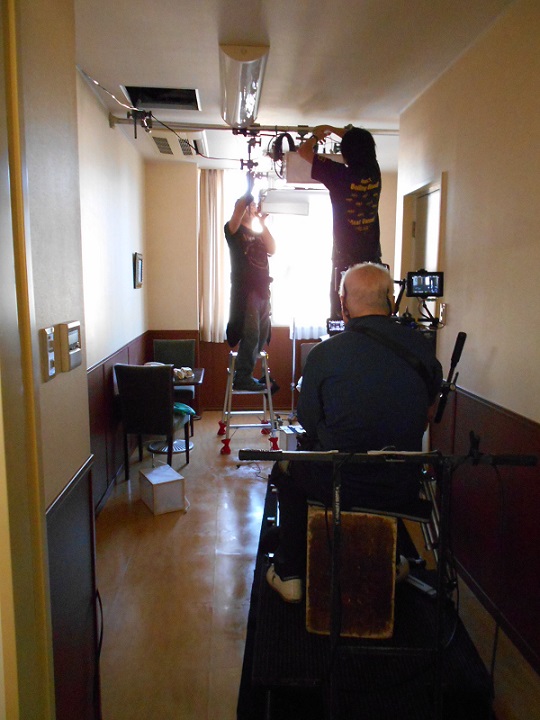 小さな個室の天井に脚立に上って撮影機材を取り付ける作業をしているスタッフの方々の写真
