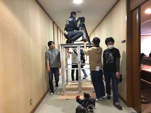 アルミで出来た人の背丈ほどの高さの作業台のような上に男性が座りカメラを設置している様子の写真