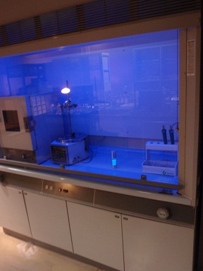 大きな1枚の透明なシャッターの奥に実験器具が置かれ、青いライトが点いたクリーンベンチの写真