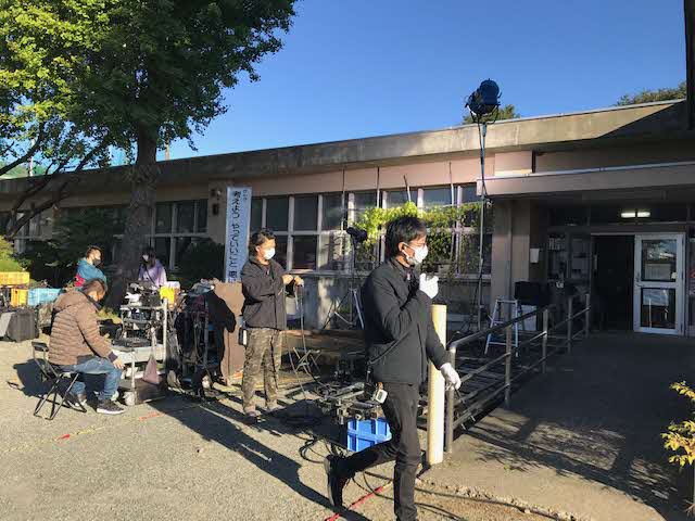 吉岡地区センター入口前の屋外に機材が置かれ、スタッフの方々が準備をしている様子の写真