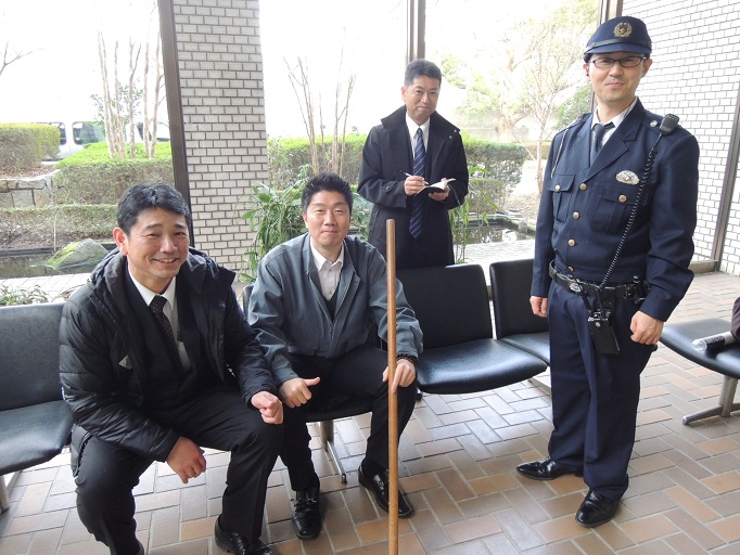刑事役の男性3名が椅子に座り、手帳を持っている刑事役の男性、警察官の制服を着た男性が写っているエキストラの方々の写真