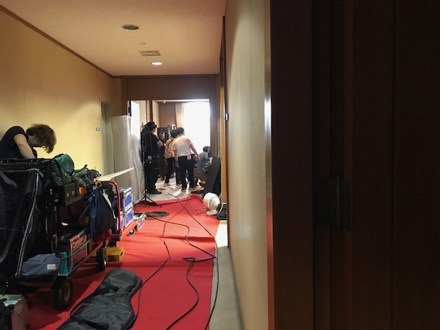 廊下に赤い布が敷かれた上に機材が置かれ、その奥の室内で撮影が行われている写真