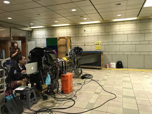 たくさんのコードがある機材の前でパソコン前に座っている男性と立っている女性の撮影現場の写真