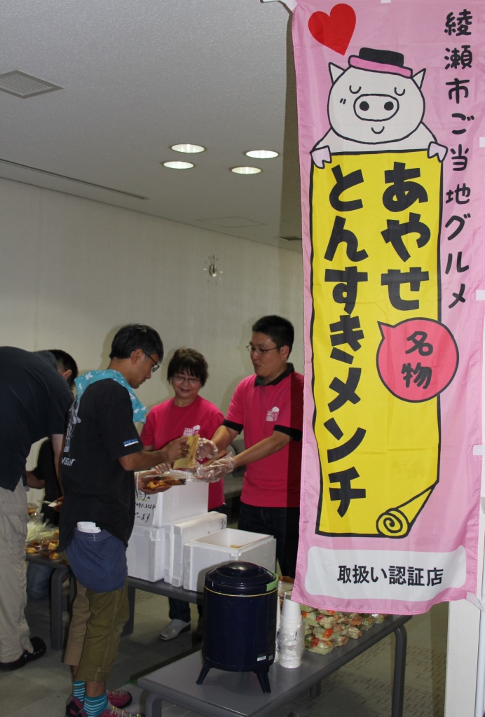 「綾瀬市ご当地グルメあやせ名物とんすきメンチ」と書かれた文字と豚のイラストがピンクの旗に描かれ、その横でピンクのシャツを着た人が男性に手渡ししている写真