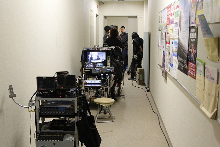 廊下に沢山の機材が準備され、その奥の部屋で撮影をしている映像が画面に映し出されている写真
