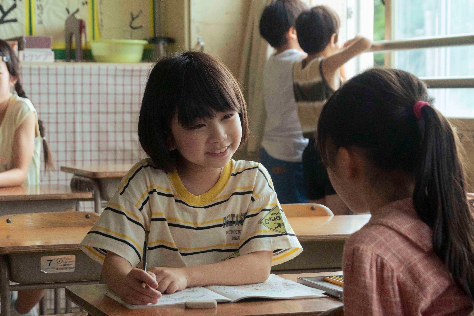 教室で机にノートを広げて座った女の子が前の席の女の子と笑顔で話をしている写真