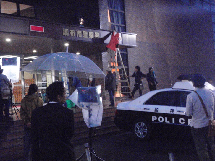 パトカーが停まっている調布南警察署入り口前で傘をさしている男性、ビニール袋が掛けられた機材、脚立に乗り警察署入り口部分で作業をしているスタッフの方がいる撮影現場の写真