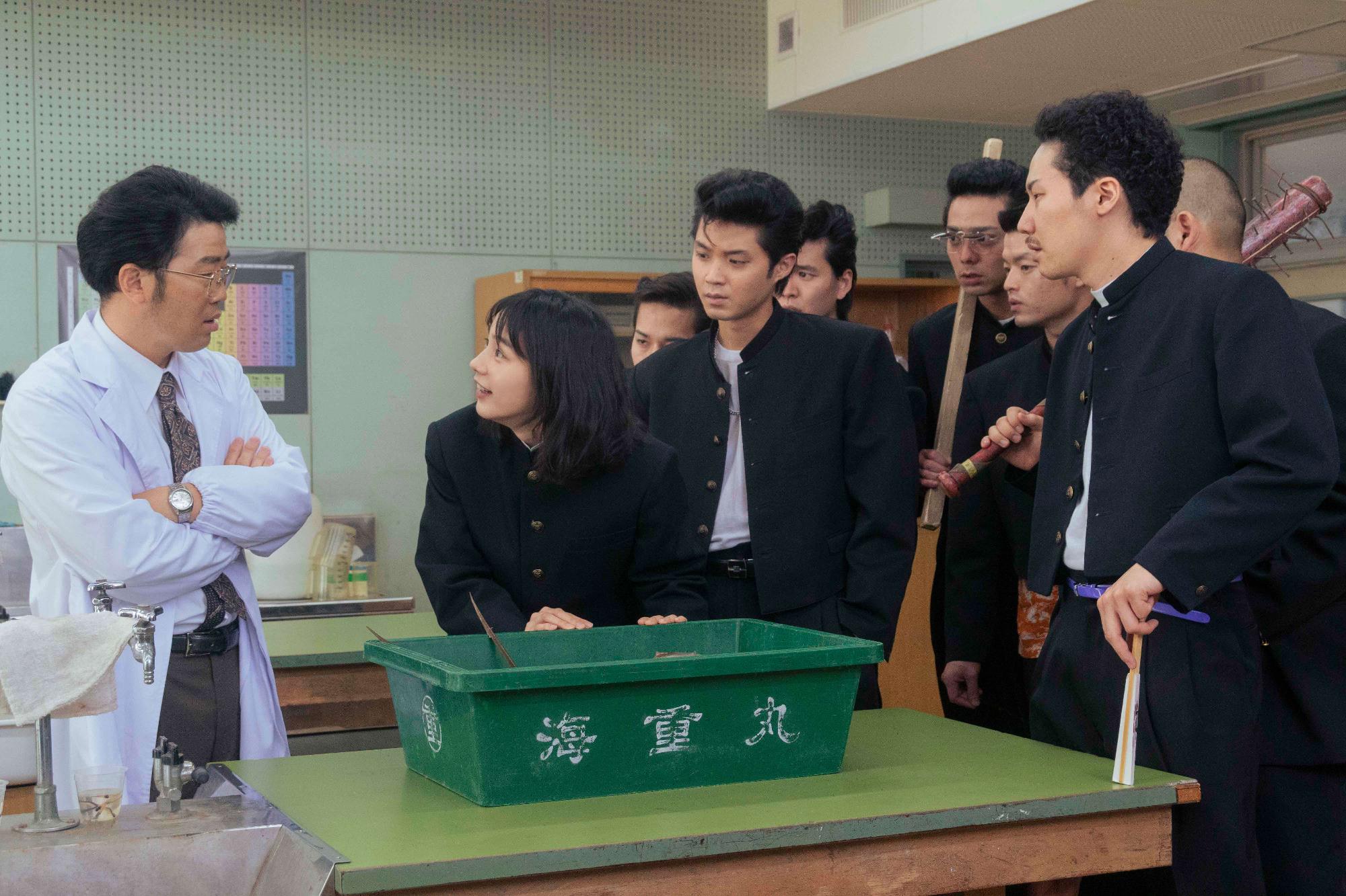 机に置かれた海重丸と書かれた緑の箱の横に白衣の腕組した男性、一人の男子学生が箱に手を置き、その後ろには学ラン姿でいろいろな道具を持った7名が立っている写真