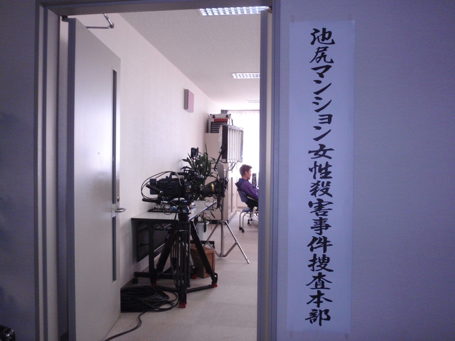 部屋の入り口の右側の壁に縦書きで「池尻マンション女性殺害事件捜査本部」と書かれた紙が貼られ、部屋の左壁側にカメラなどの機材が置かれている写真