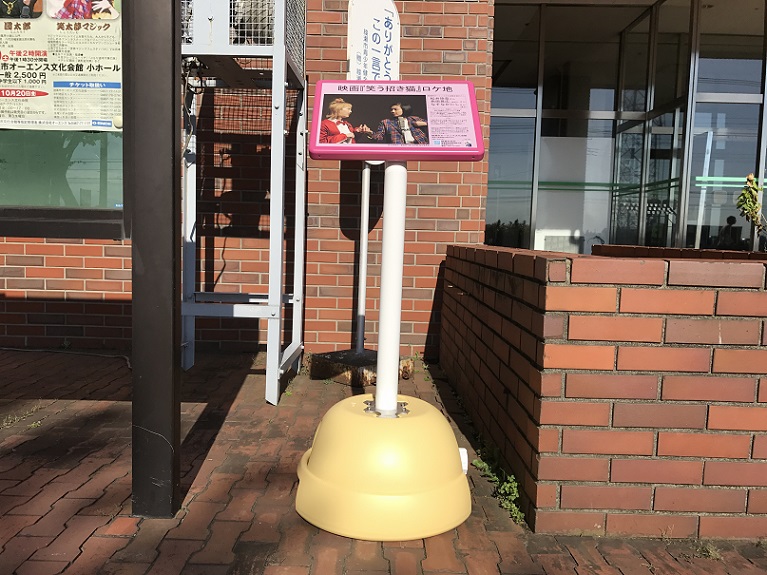 綾瀬市オーエンス文化会館入り口付近の掲示板の横にロケ地看板が置かれている写真