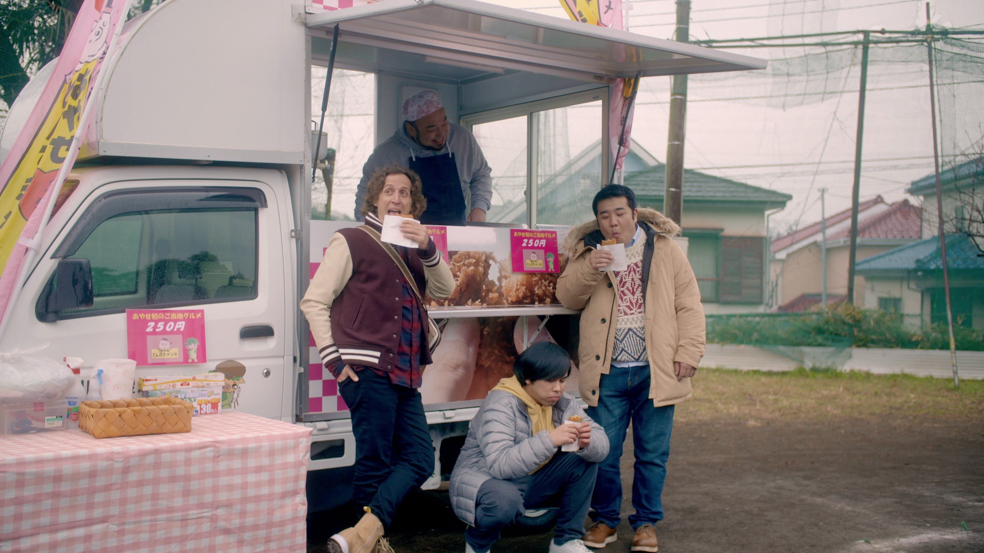 軽トラックキッチンカーの前で揚げ物を食べている堤下敦さん他3名の役者さんが写っている映画ルーツのワンシーンの写真
