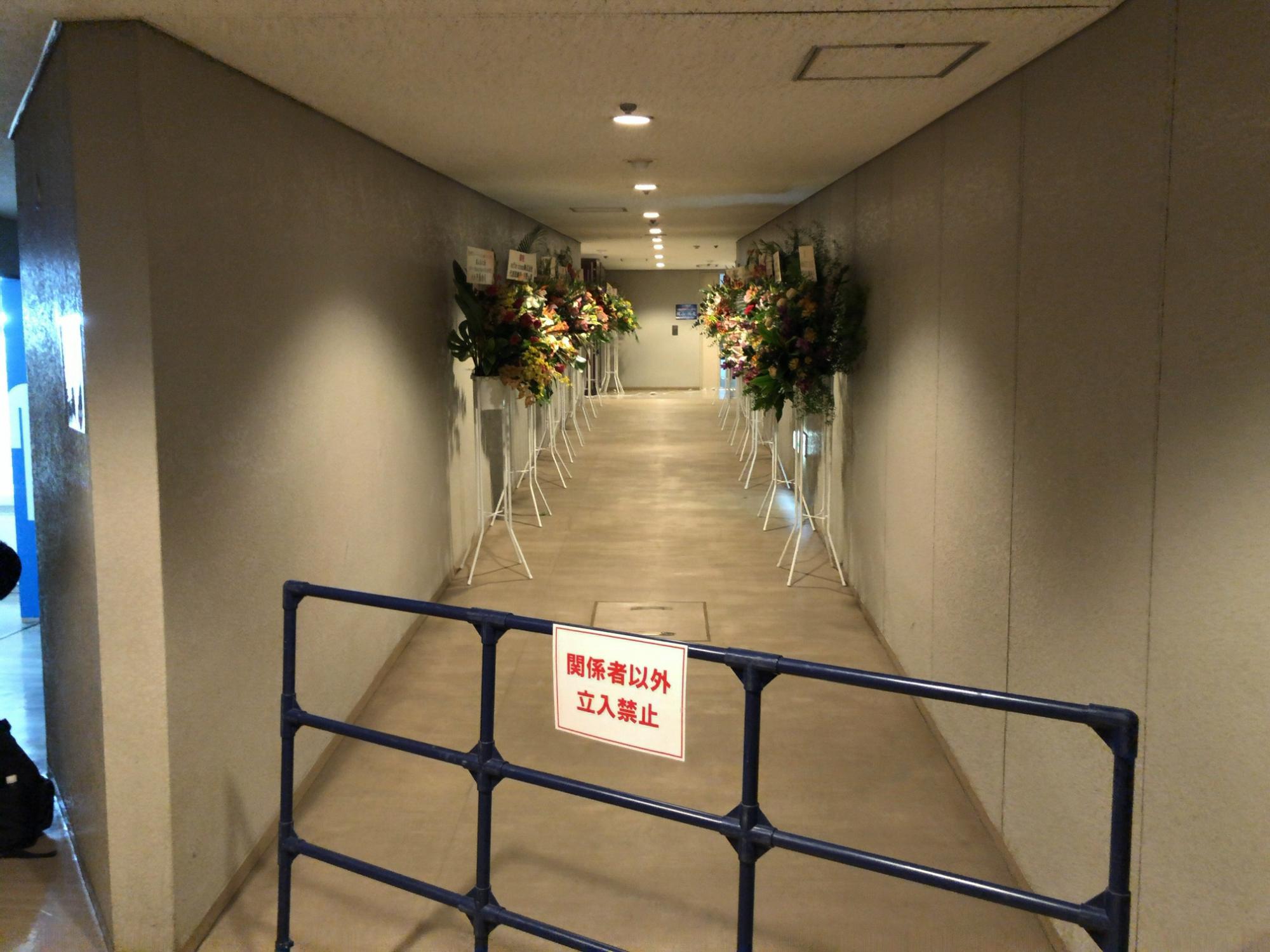 通路の左右にスタンド花が間隔をあけて奥まで並び、手前の柵に「関係者以外立入禁止」の札がついている写真