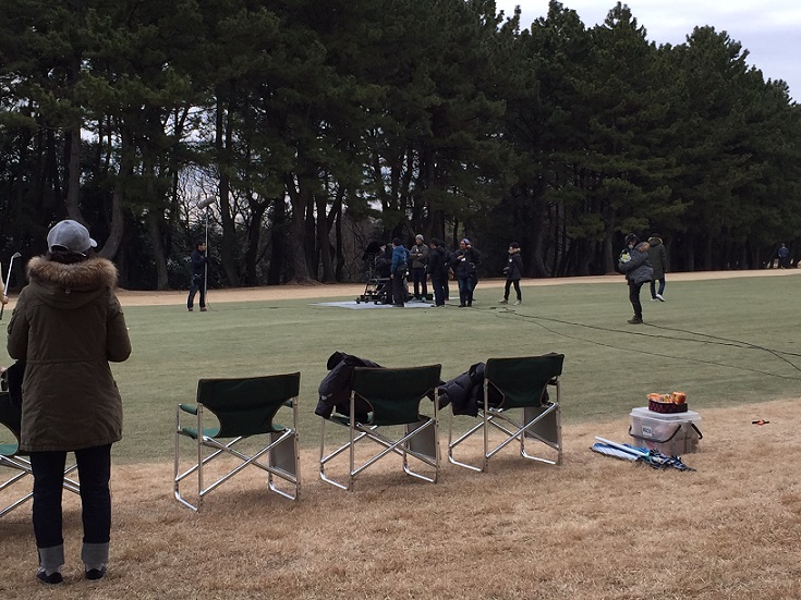 ゴルフ場のコース中央で撮影が行われており、離れた場所にアウトドア用の椅子が並び、椅子の横に撮影の様子を見ている人が写っている撮影風景の写真