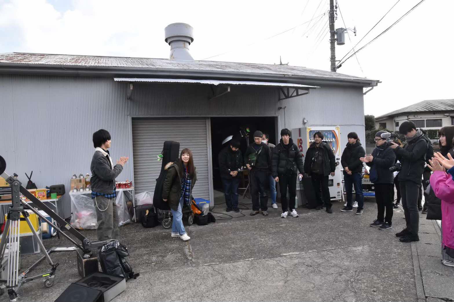 ブタッコリーのマスコットチェーンがついたギターケースを背負った村田寛奈さんが倉庫の前でスタッフの方々に拍手されている写真
