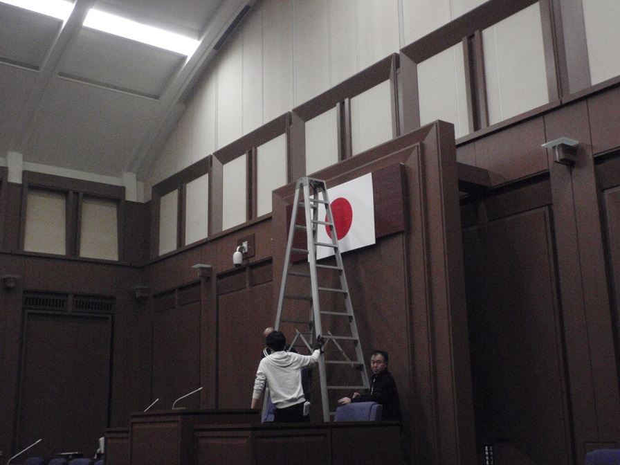 日の丸の国旗が議長席の後方の壁に掲示され、その前に脚立が置かれスタッフの方々が撮影の準備をしている様子の写真