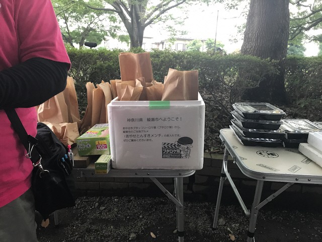 「神奈川県綾瀬市へようこそ！」と書かれた貼り紙が簡易テーブルの上に置かれた発泡スチロールに貼られ、中に沢山のメンチカツが紙袋に入っている写真