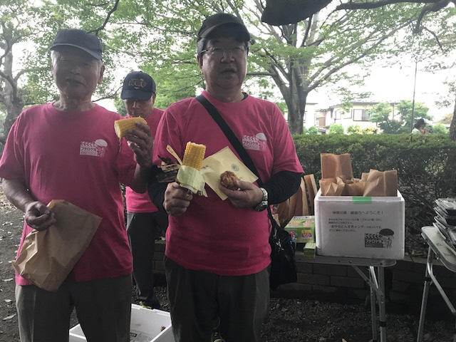 ピンク色のシャツを着た3名の男性スタッフがメンチカツとトウモロコシを食べている写真