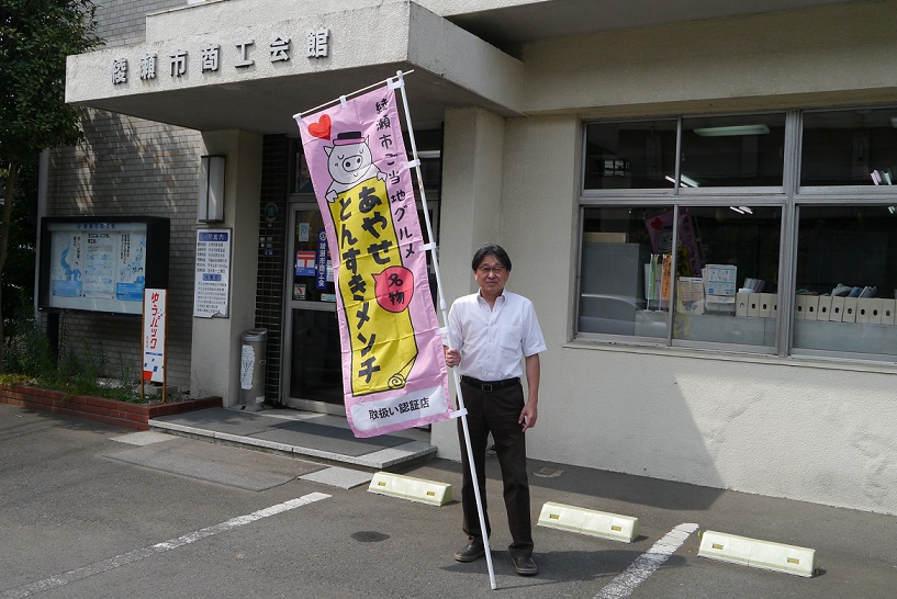 商工会館の入り口の前で男性があやせとんすきメンチと書かれたのぼり旗を持って立っている写真