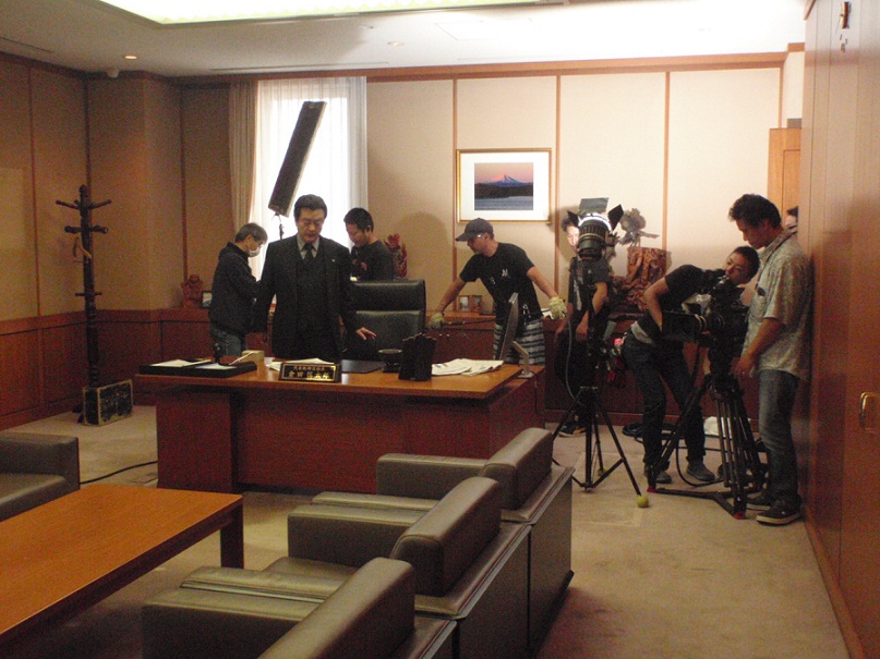 大きな机の前で坐ろうとしている俳優さんと、撮影の準備をしているスタッフやカメラマンの人達の写真