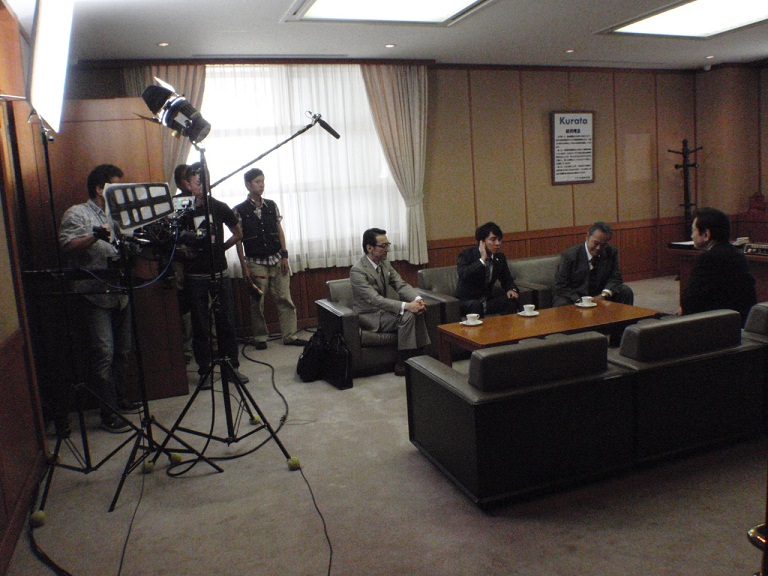 大きな机を中央にソファーに座って演技をしている4名の俳優さんを、音声さんが長いマイクで声をひろっている写真