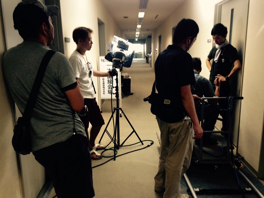 廊下に照明器具が設置され、5名の撮影スタッフが映像を見ている撮影風景の写真