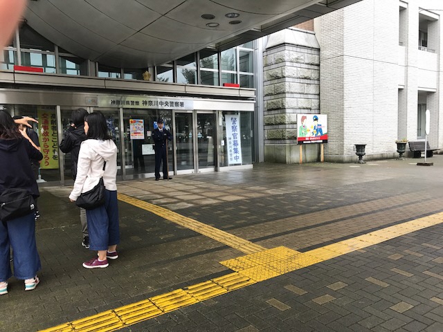 正面に「神奈川中央警察署」と書かれた建物の前に制服姿の警察官が立ち、手前左側にスタッフが立っていて撮影が行われている現場の写真