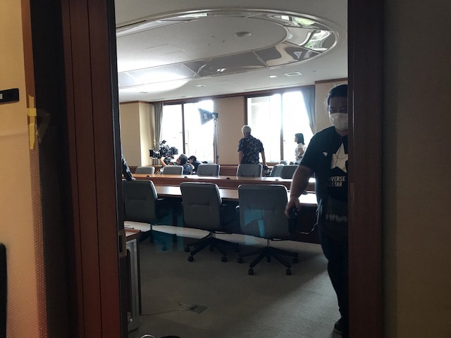 広い会議室の奥に設置された照明の下でカメラを構えているスタッフが撮影を行っている写真