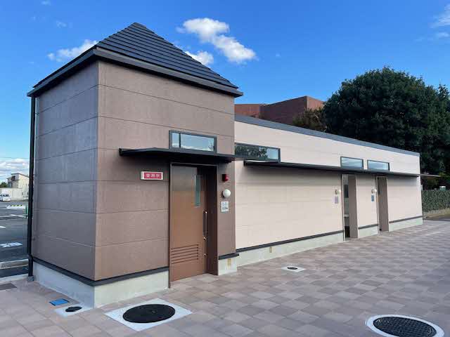 茶色の外壁で屋根が三角屋根になっているバリアフリートイレと横にベージュ色の外壁の女性用と男性用のトイレの施設が設置されている防災トイレの外観写真