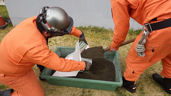 スコップを使い、容器に入った土を土のう袋に入れる隊員の写真
