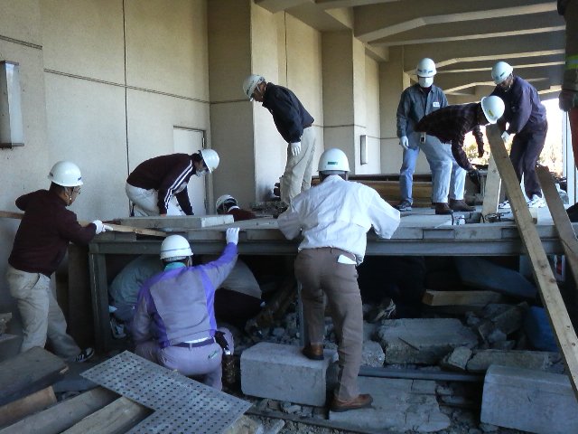ヘルメット姿の人達がコンクリートの上や横、下に入って神奈川県防災センターの派遣研修に参加している写真