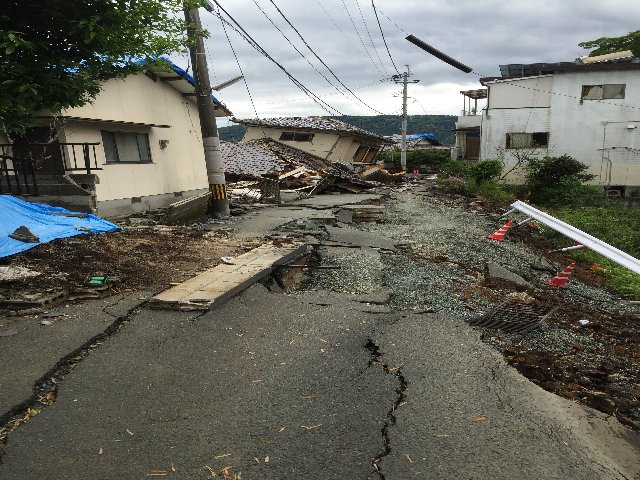 表面にひびが入り崩れている道路、奥には家が倒壊している写真