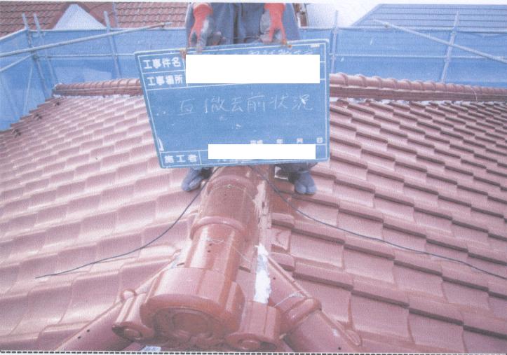 茶色の瓦屋根の上で作業員が「瓦撤去前状況」と書かれたパネルを持っている写真