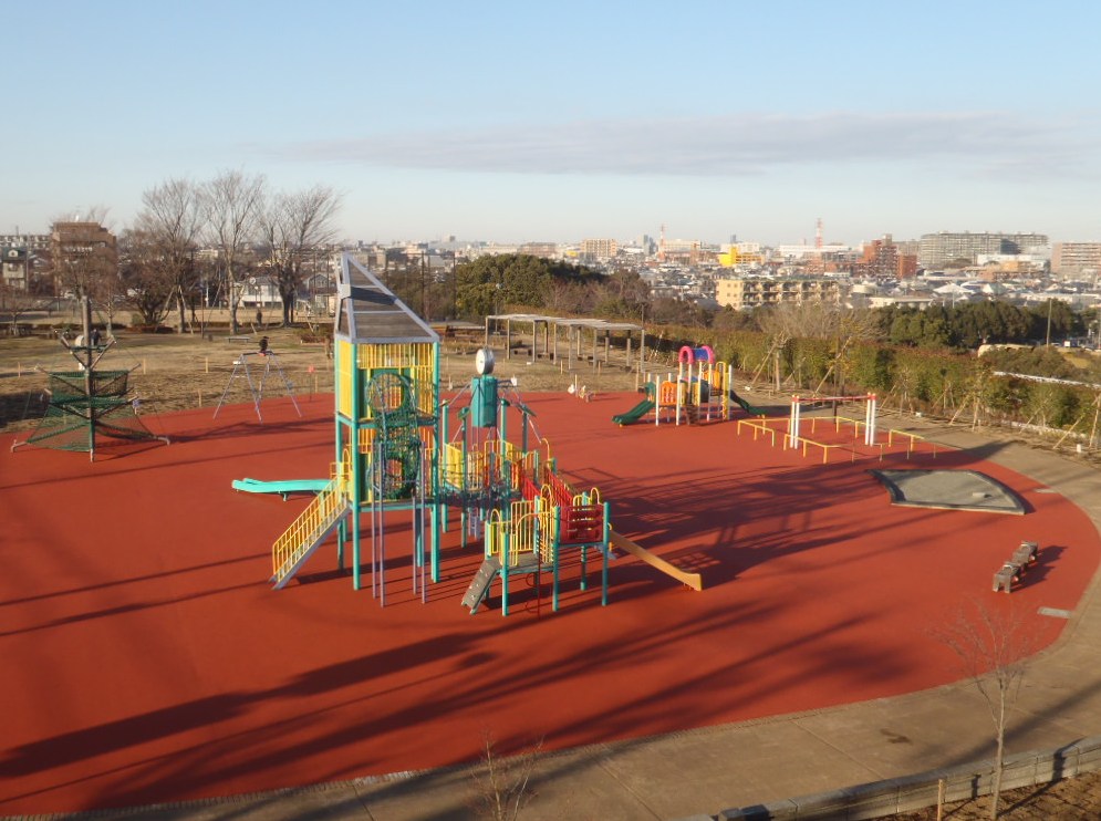 茶色のマットが敷かれ砂場やベンチ大小2つの複合遊具などがある遊具広場を全体が写るよう上から撮影した写真
