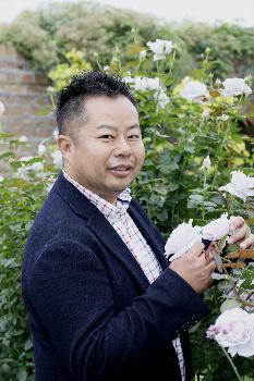 チェック柄のシャツにネイビーのジャケットを着て、白いバラの花を持ちこちらを向いている河合伸志氏の写真