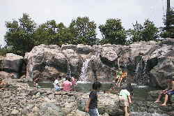 大きな岩の岩肌から流れる水が溜まっている場所で水遊びをする子供たちの写真