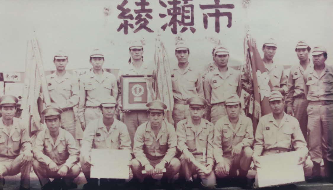 綾瀬市の文字を背景に優勝旗などを持って写っている綾西分団の方々の白黒写真