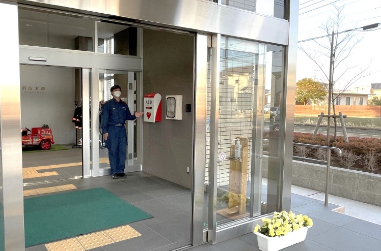消防本部自動ドアの入口奥の右側に設置されているAEDと非常電話の位置を示している男性隊員の写真