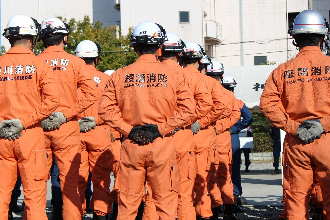 ヘルメットを被り背中に「綾瀬消防」と書かれたオレンジ色の活動服を着た方々を後ろから撮影した写真