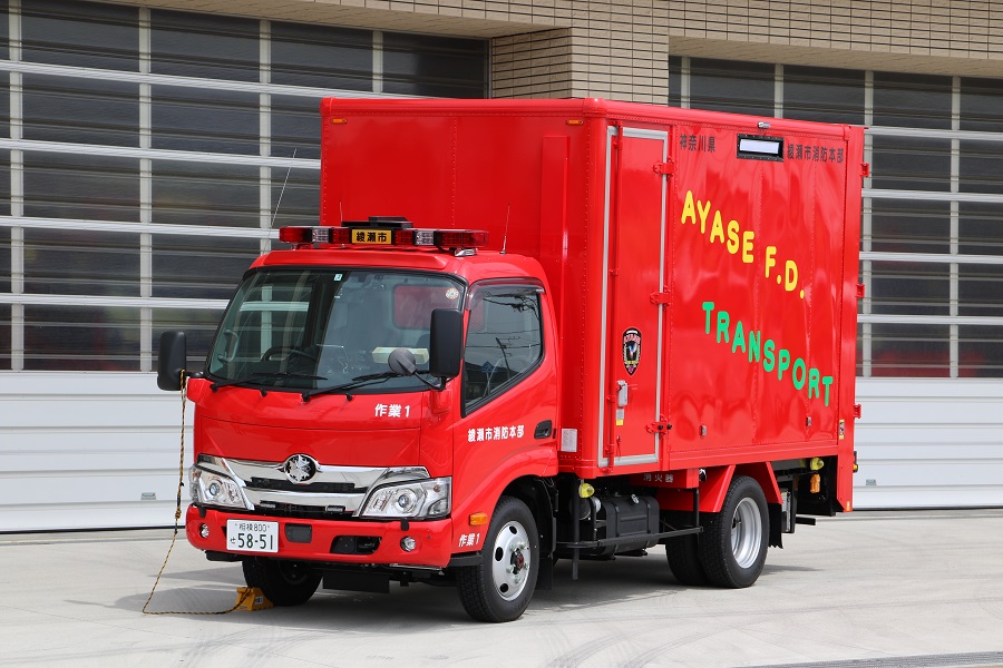 赤い車体の大きな荷台の側面に黄色で「AYASE F.D.」緑色で「TRANSPORT」と斜めに書かれている資器材搬入車の写真